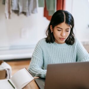 Una mujer joven revisa su computador portátil con un cuaderno abierto a un lado, anotando lo que encuentra: es su chequeo por cuenta propia